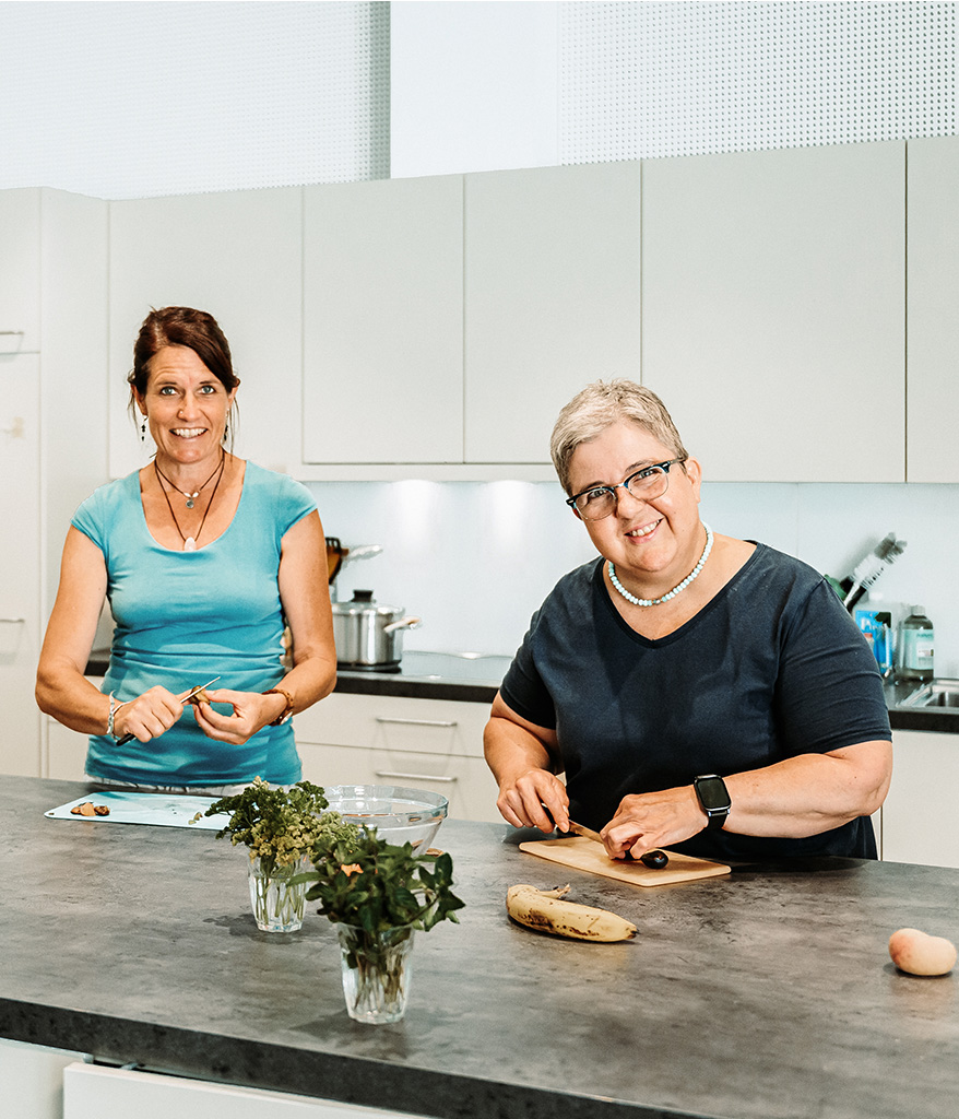 Eine Frau mit blauem T-Shirt und eine Frau mit schwarzem T-Shirt schneiden in der Küche Früchte.