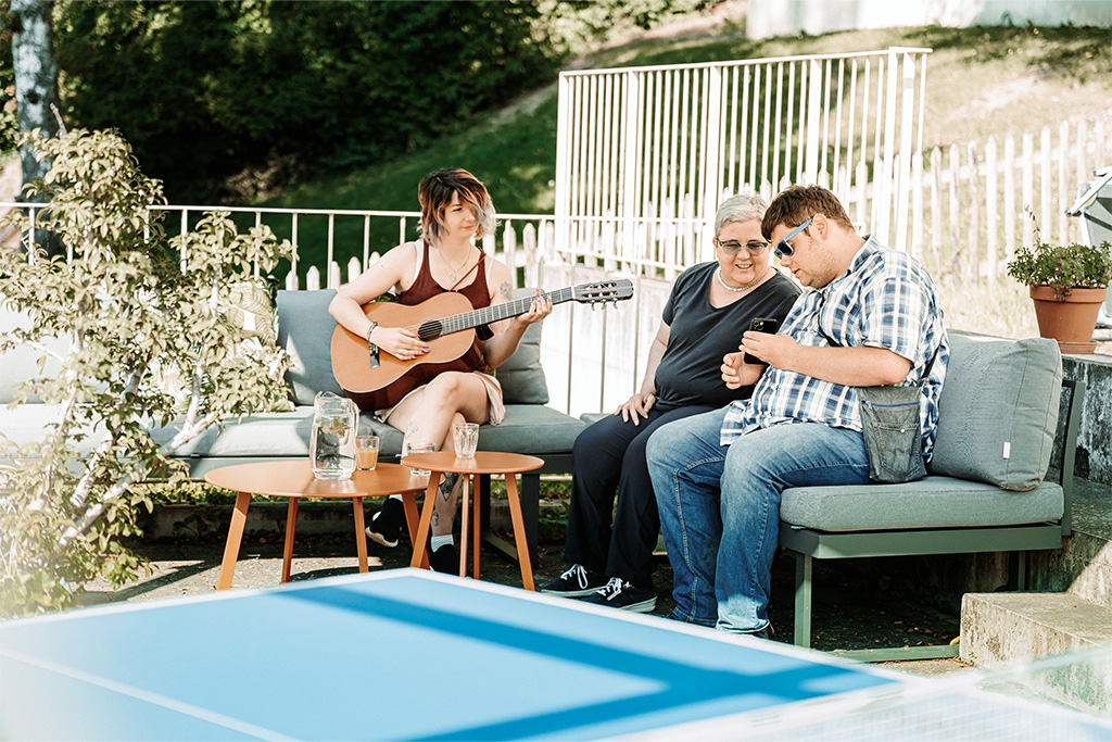 3 Personen sitzen draussen auf einem Sofa, eine davon spielt Gitarre