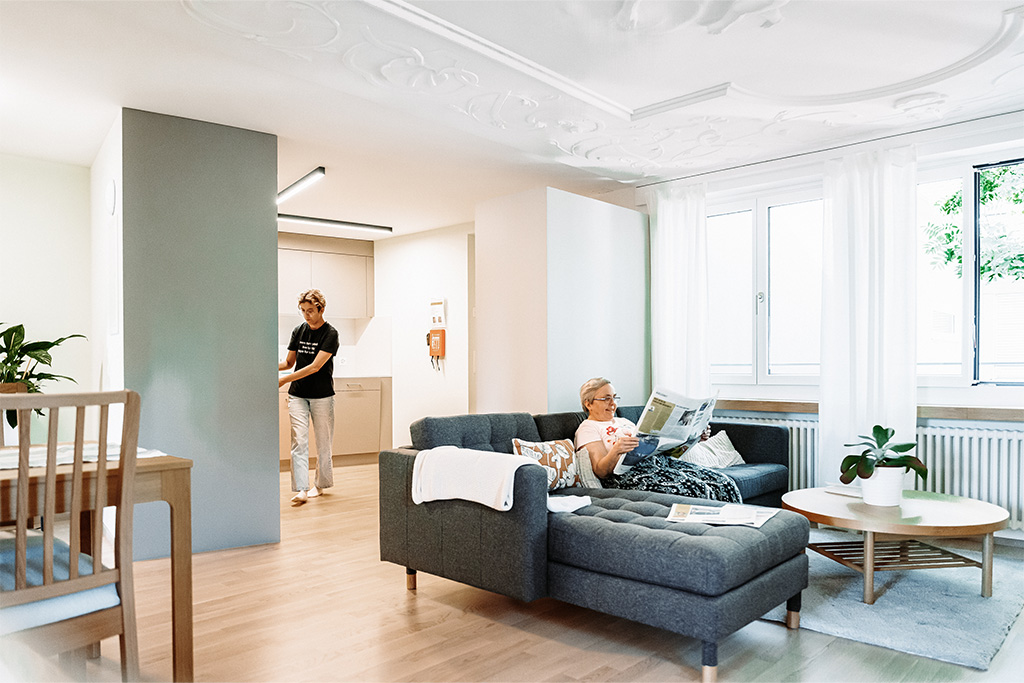 Wohnstube mit Tisch, Stühlen und schwarzem Sofa. Eine Bewohnerin ist in der Küche und eine sitzt auf dem Sofa und liest Zeitung
