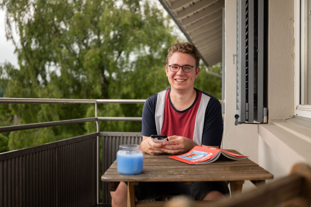 A young man is sitting at a wooden table on the balcony. There is a book and a candle on the table. He is holding a cell phone in his hand.