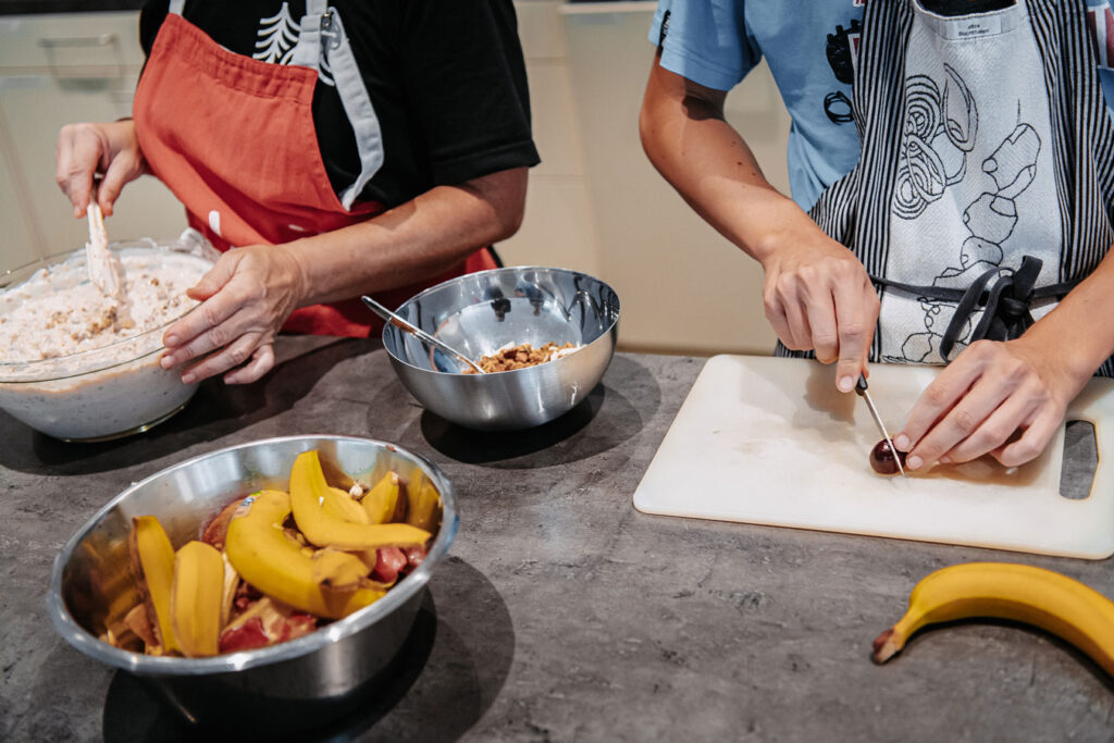 Two people with cooking aprons in the kitchen. One person is mixing fruit with yogurt to make a muesli. The other person is cutting fruit.