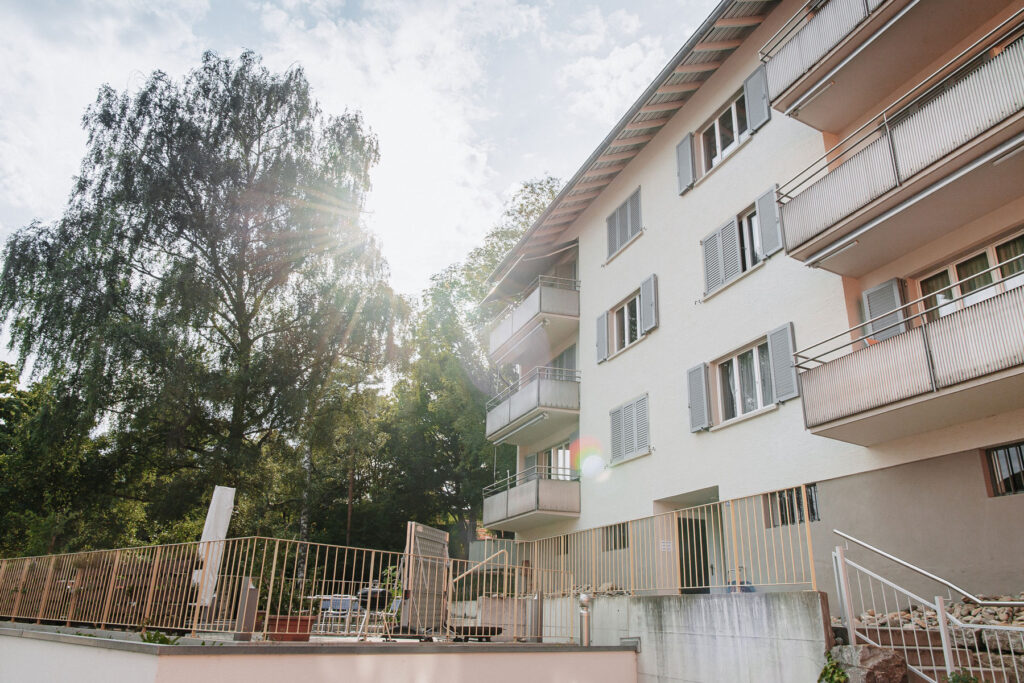 Die Fassade und der Aussensitzplatz eines Hauses ist zu sehen. Auf dem Sitzplatz befinden sich Sonnenschirme und ein Tischtennis-Tisch.