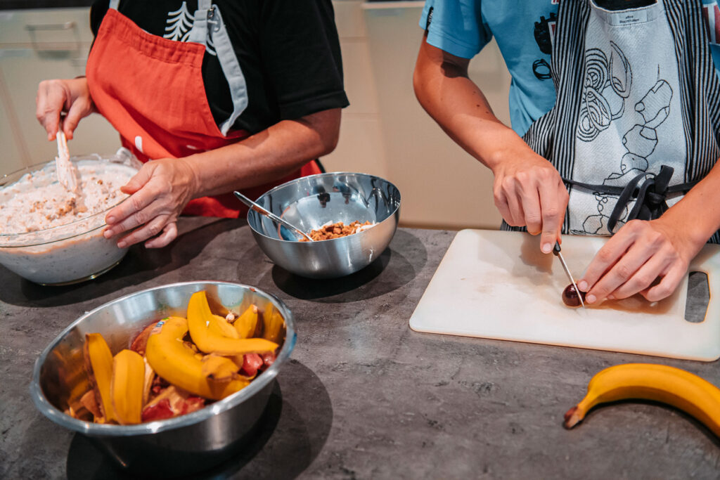 Two people with cooking aprons in the kitchen. One person is mixing fruit with yogurt to make a muesli. The other person is cutting fruit.