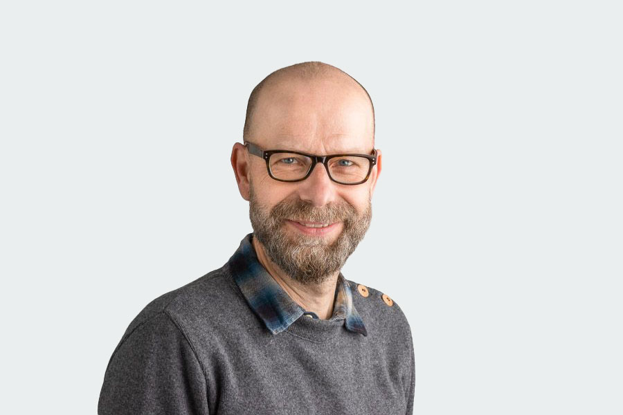 Jens Förster, job coach and contact person for our employees and external companies when it comes to inclusive workplaces. He wears glasses, a full beard, a multicolored shirt and a grey sweater.