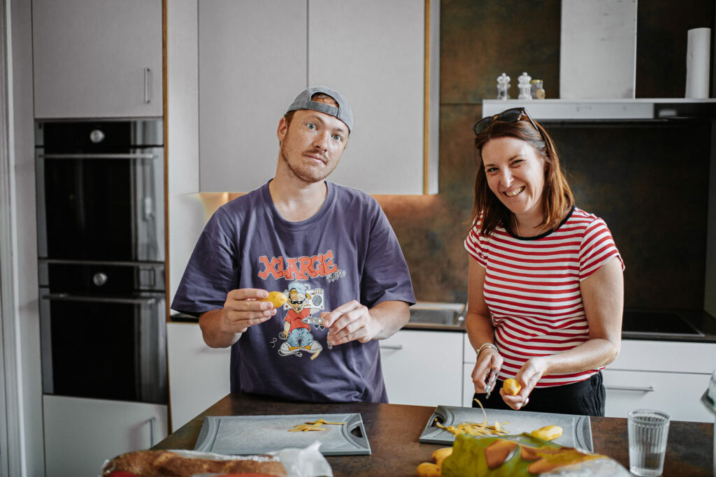 A man and a woman are preparing potatoes in a kitchen. Both are looking into the camera.