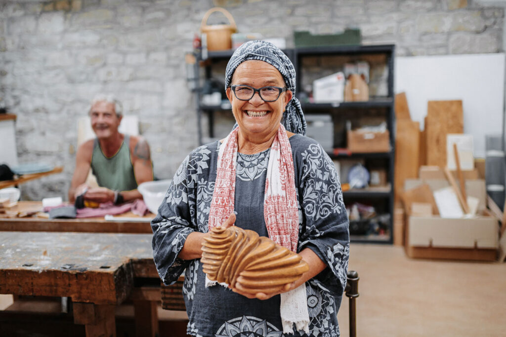 In the foreground, a woman with a large soapstone in her hand is beaming. She is in a creative room. In the background sits a second person who is doing grinding work by hand.