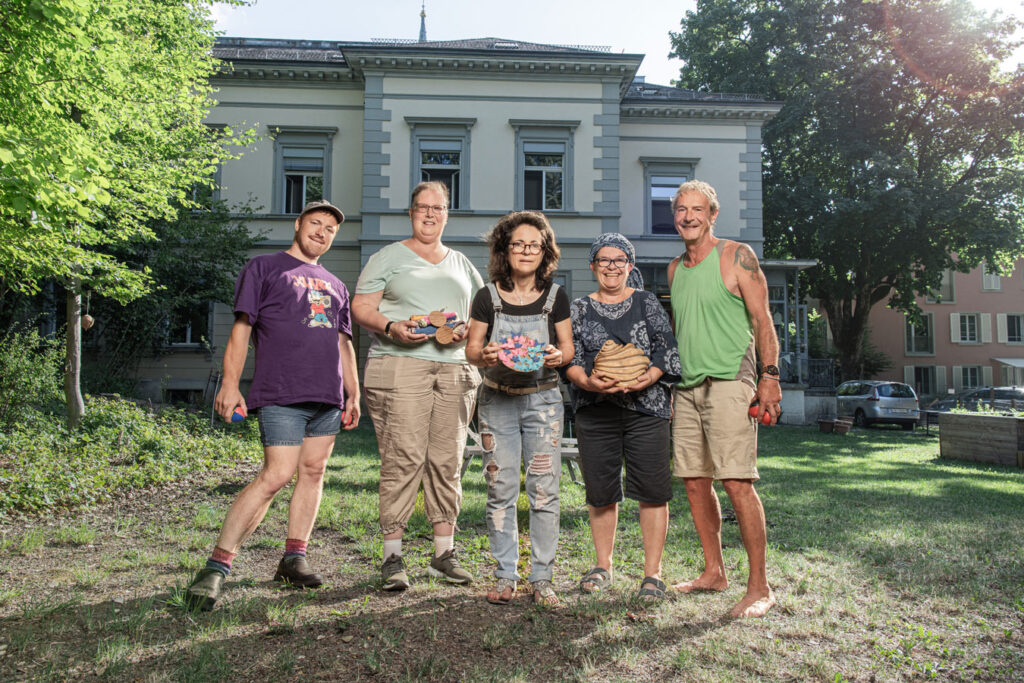 Five people of different ages are standing in the garden in front of a house. They are all holding an object in their hands that they either like to play with or have made themselves.