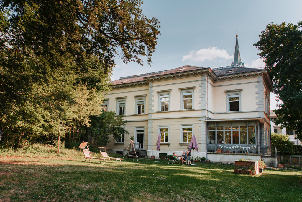 Villa in Schaffhausen. Grosser Garten mit vielen verschiedenen Sitz-Gelegenheiten vor einem Jugendstil-Haus mit vielen Fenstern und einer dezent crèmefarbigen Fassade.