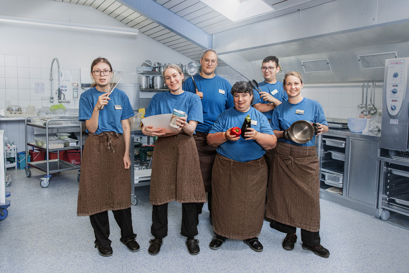 Sechs Personen des Teams Bistretto. Sie befinden sich in der Küche und haben alle einen Gegenstand in der Hand, zum Beispiel Kochtopf, Schöpflöffel, Schwingbesen.