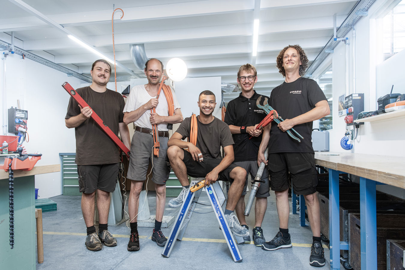 Picture with 5 people from the Engineering and Maintenance team in a workshop room. Each person is holding an object: spirit level, a luminous lamp, cables and two different pliers.