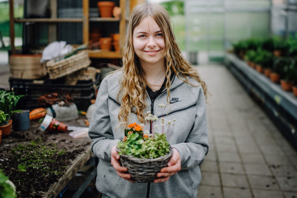 Junge Frau mit grauer Trainingsjacke hält ein Korb mit Blumen in der Hand.