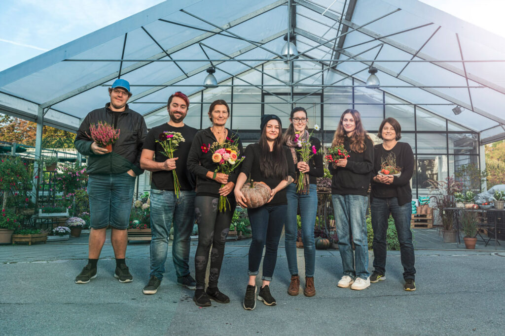 Seven people who work in the nursery are standing in front of the flower store. They are carrying various flowers in their hands.