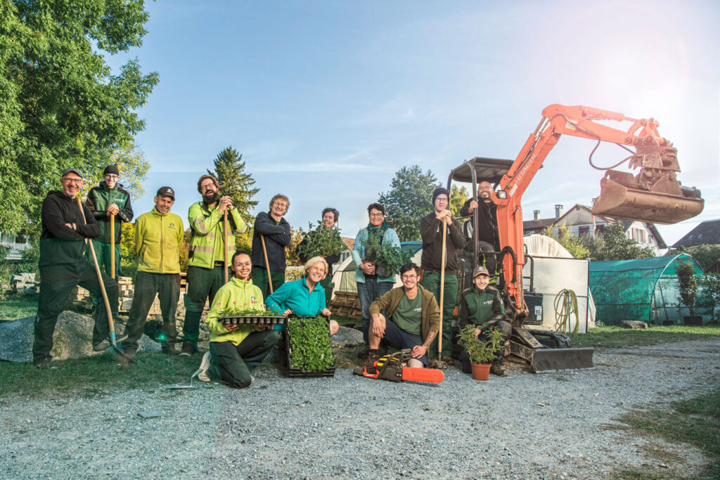 Thirteen people working in horticulture are standing and sitting on a gravel parking lot. An excavator, plants and various tools are visible.