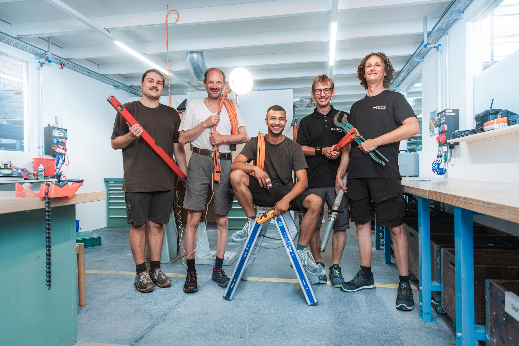 Picture of 5 people from the Engineering and Maintenance team in a workshop room. Each holds an object in his hand: spirit level, a luminous lamp, cables and two different pliers.