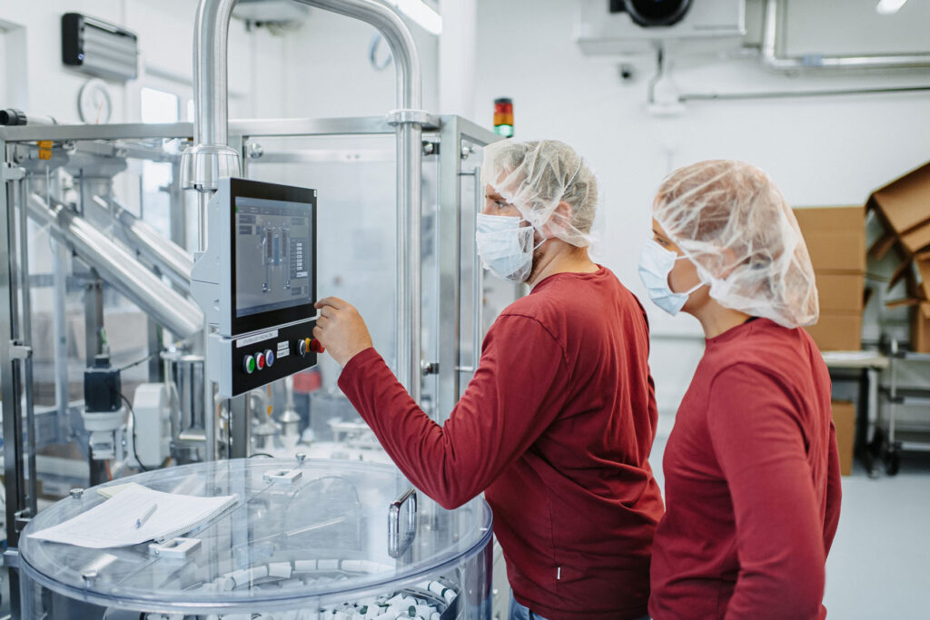 Two employees from the food team are in a production room. Both are wearing a dark red long-sleeved shirt, face masks and a hood. The employee is operating the electronic controls of a food filling machine.