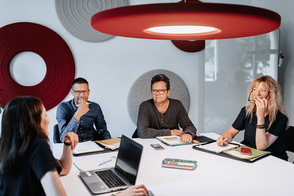 Bild von 4 Personen des Fachbereichs Mitarbeitende während einer Teamsitzung. Sie diskutieren an an einem weissen Tisch und machen sich Notizen auf einem Block und mittels Laptop.