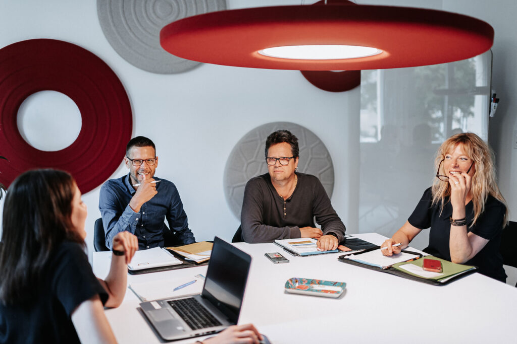 Four people are sitting at a table. They are discussing and taking notes on a notepad and laptop.