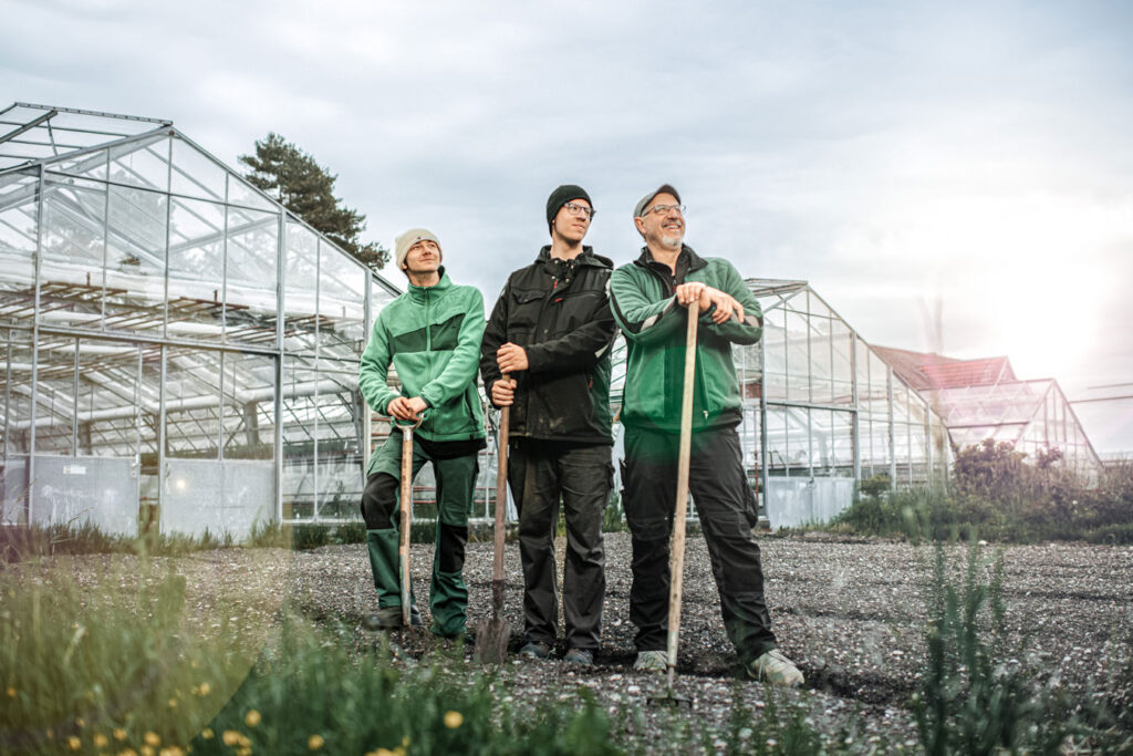 Three people stand in a field in front of greenhouses and look up at the sky. They are holding gardening tools in their hands.