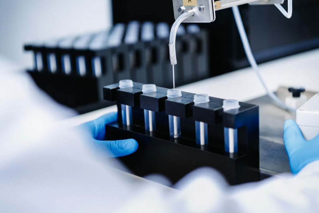 A filling device from which liquid is filled into a reagent tube. The tube is placed in a block with a total of 5 tubes. A person wearing blue nitrile gloves holds the block.