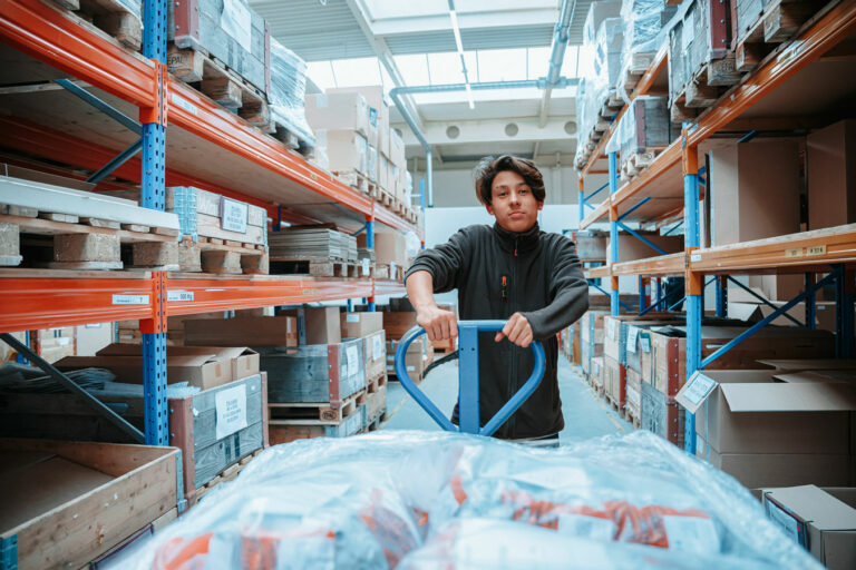Young man transports goods with the pallet trolley and stands in the high-bay warehouse.