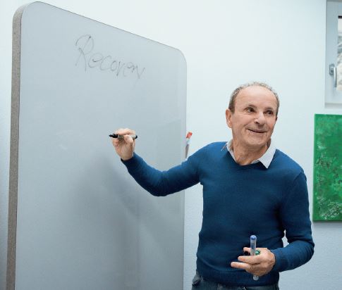 Ein Mann mit blauem Pullover steht mit einem Stift in der Hand vor einer weissen Tafel.