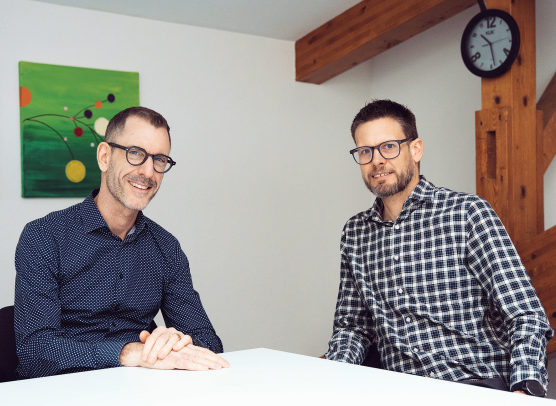 Zwei Männer sitzen an einem Tisch, beide tragen ein Hemd und eine Brille.